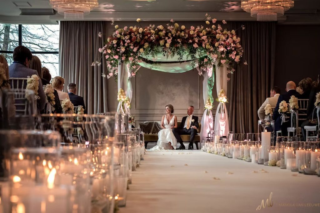 Bruidspaar zit onder een bloemenboog tijdens de ceremonie, met gasten en kaarsen langs het gangpad