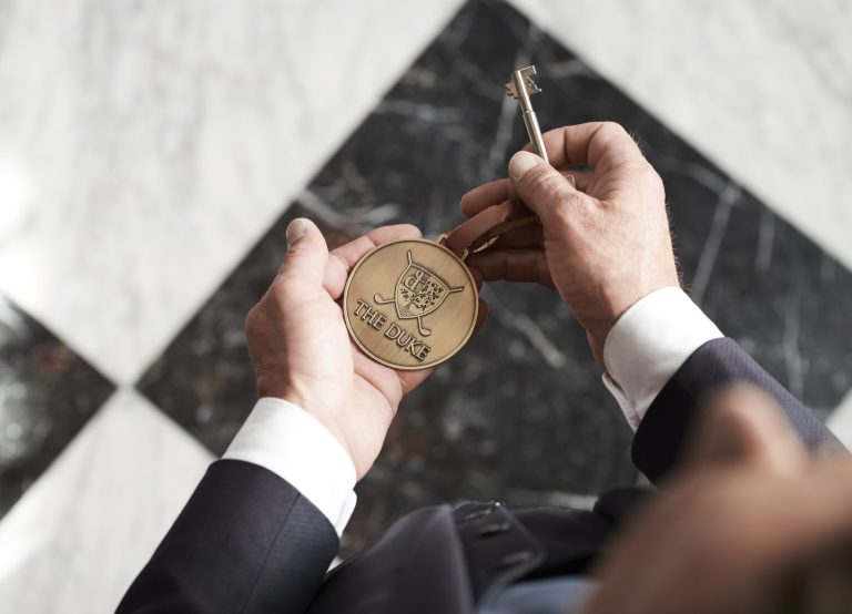 Handen houden een sleutel met ronde hanger met het opschrift THE DUKE boven een zwart-witte tegelvloer