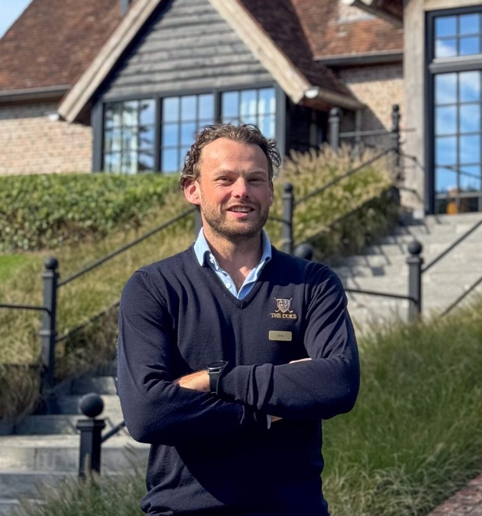 Man in een trui met The Duke-logo staat met gekruiste armen voor een gebouw met een buitentrap