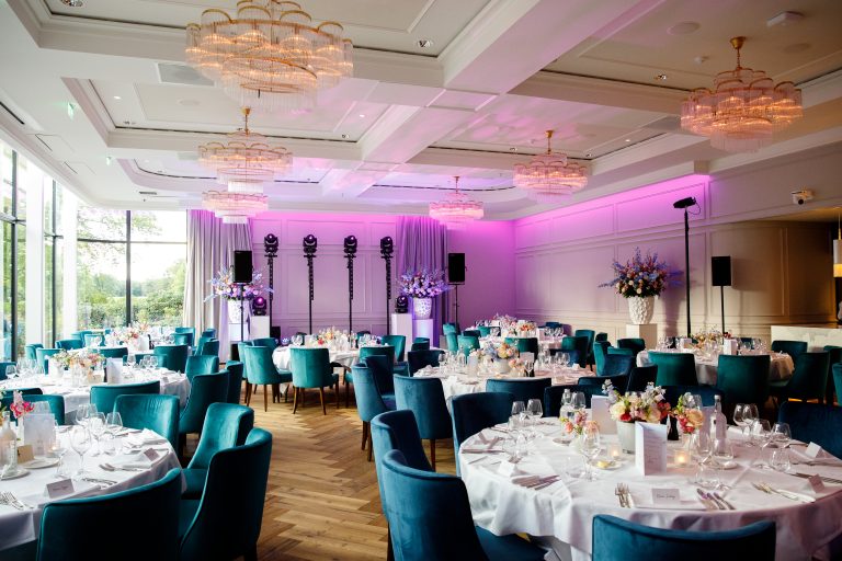 Feestzaal bij The Duke met gedekte ronde tafels, blauwgroene stoelen, kroonluchters en roze verlichting