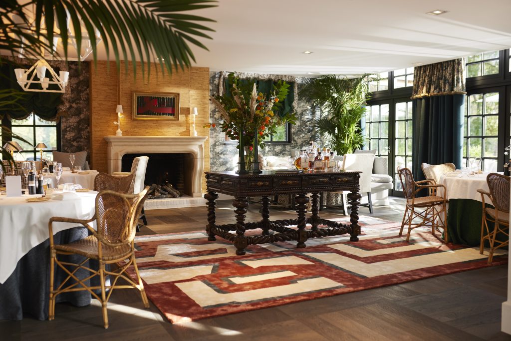 Gedekte tafels en rieten stoelen rond een houten tafel met bloemen, bij een open haard en hoge ramen