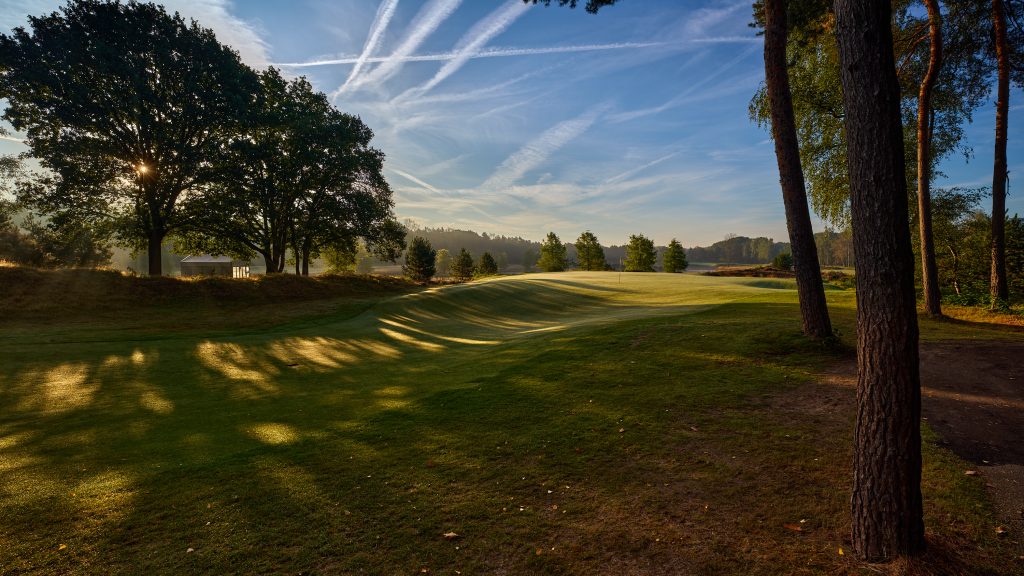 Hole 16 op de golfbaan van The Duke, met lange boomschaduwen over het gras en laagstaande zon tussen de bomen