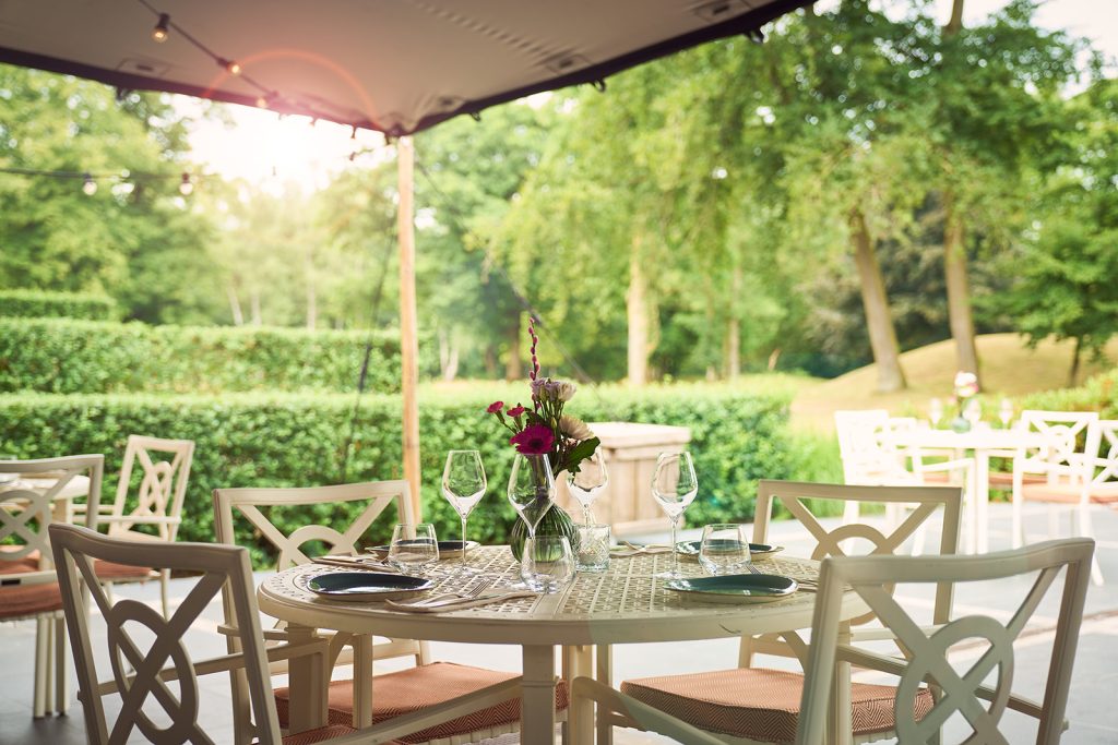 Gedekte ronde tafel met bloemen en wijnglazen op het overdekte terras van The Courtyard, omringd door groen