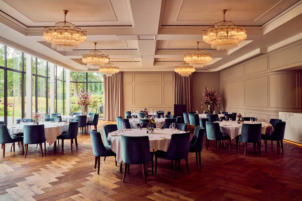 Gedekte ronde tafels met groene stoelen, bloemen en kroonluchters in een zaal met grote ramen bij The Duke