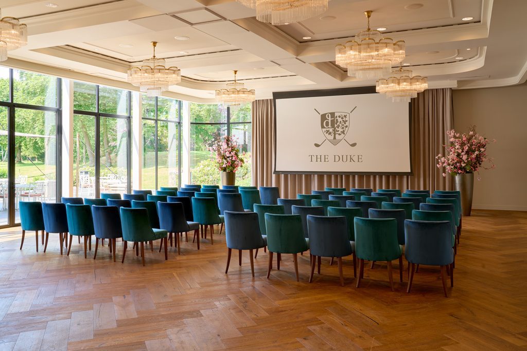 Zaal met rijen blauwe en groene stoelen, kroonluchters en een projectiescherm met het logo en de tekst THE DUKE