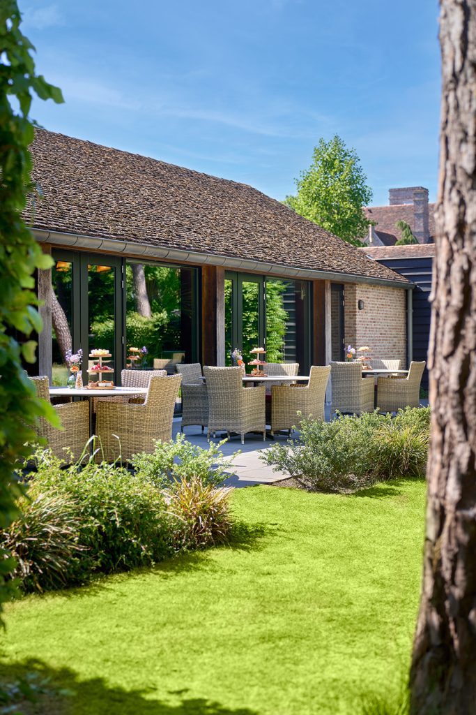 Terras van The Lodge met gevlochten stoelen en tafels met etagères, omringd door een groene tuin