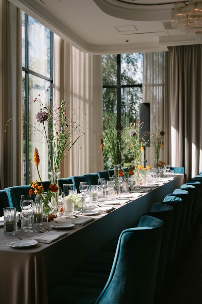Lange gedekte tafel met hoge bloemenvazen en blauwgroene stoelen in een zaal met grote ramen