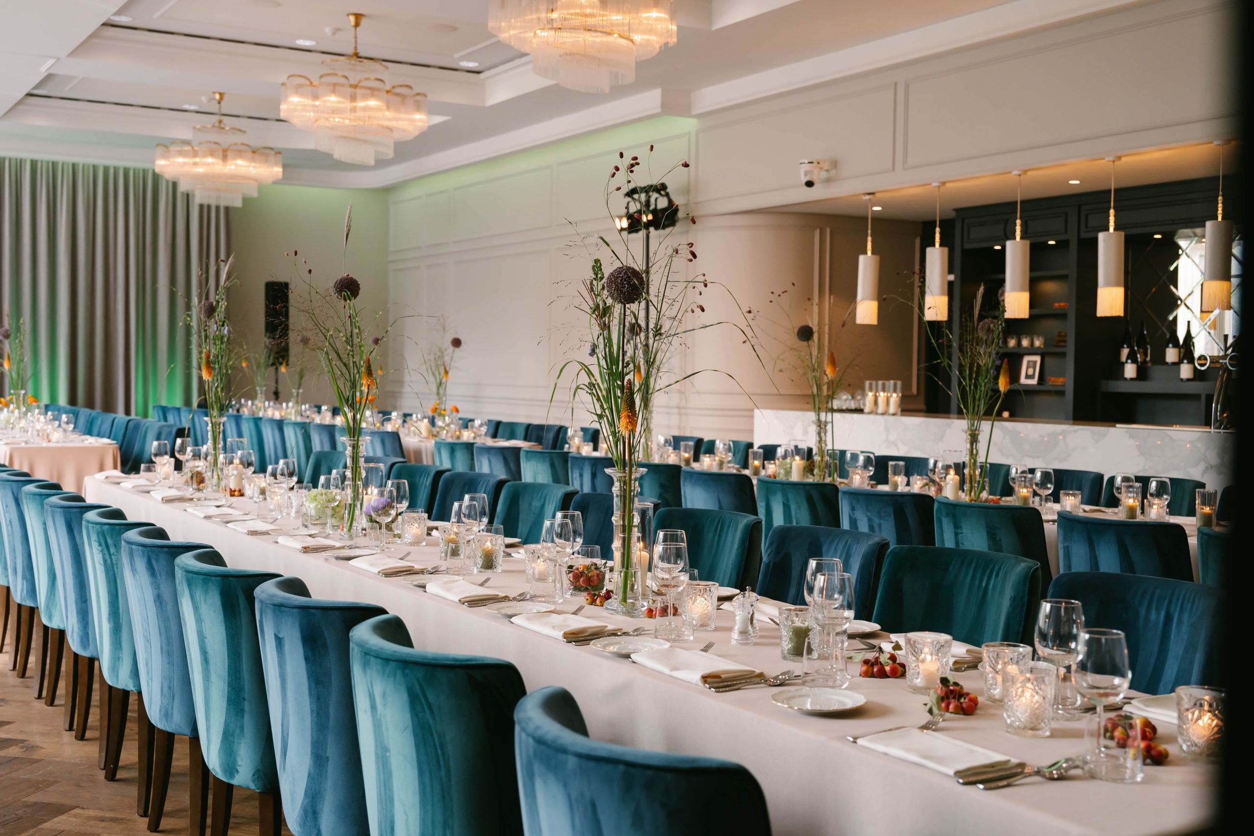 Lange gedekte tafels met hoge bloemstukken en blauwgroene stoelen in een zaal met kroonluchters