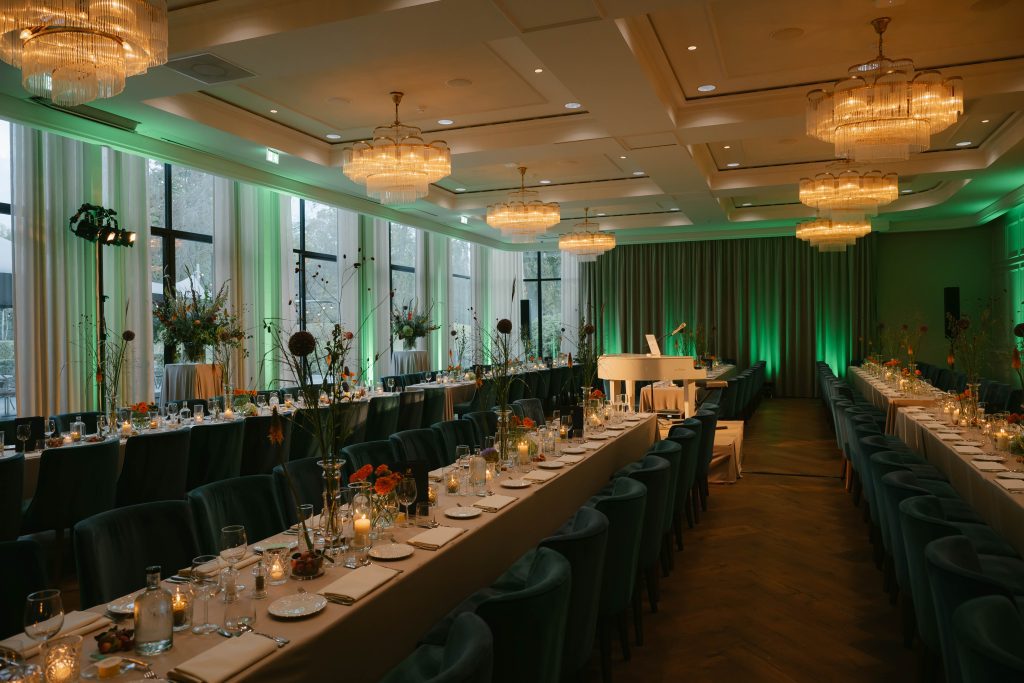 Lange gedekte tafels met bloemen en kaarsen in een zaal met kroonluchters, groene verlichting en een witte vleugel
